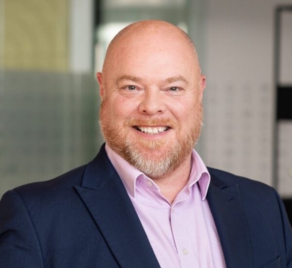 A professional portrait of a man with a bear smiling, wearing a dark suit jacket and a light pink shirt.