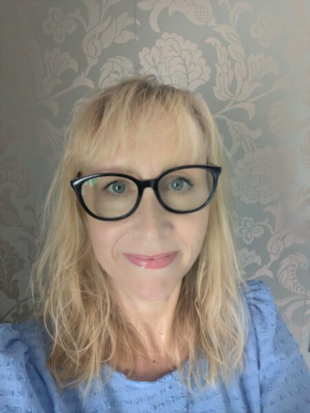 A portrait of a women with long blonde hair. She has thick black framed glasses and is wearing a blue top.