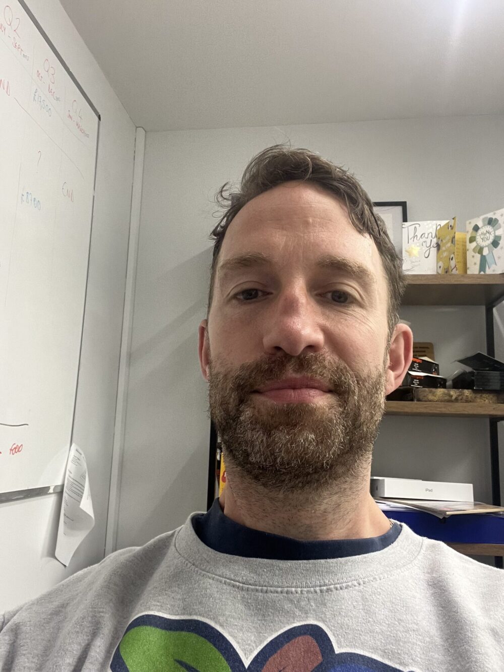 A picture of a man with short brown hair and a beard, He is wearing a light coloured shirt.