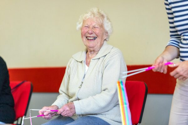 An elderly woman with white hair, smiling and holding a colourful ribbon