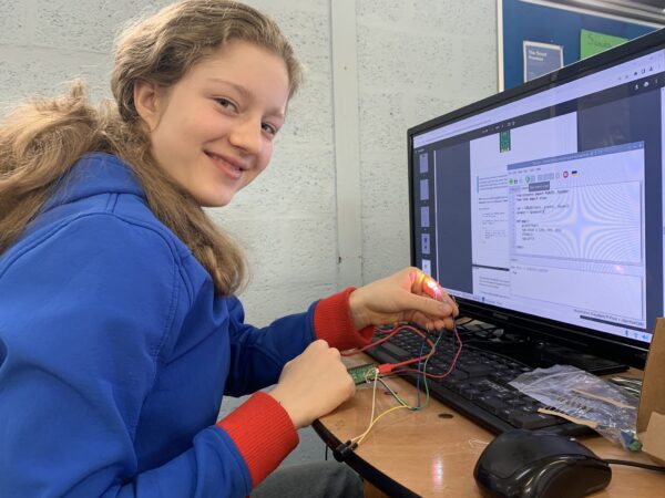 A young girl seated at a desk holding some colourful wires connected to a computer