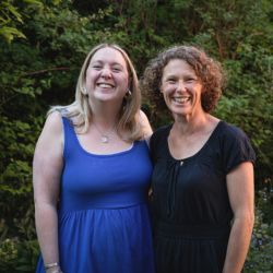 Two women smiling at the camera, one is wearing a blue dress and the other a black dress.