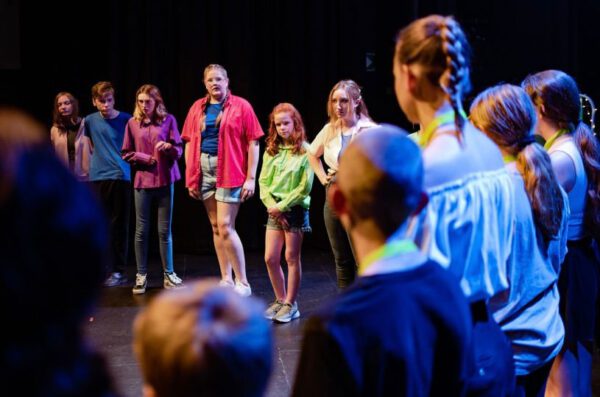A group of children and young teenagers, standing together on a stage under dramatic lighting.