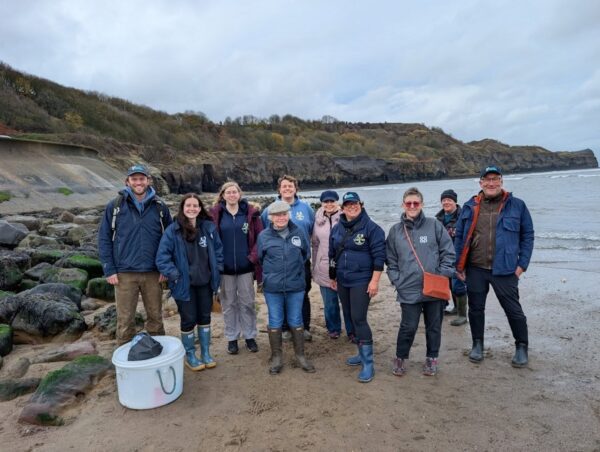 About 10 people standing on a beach or shoreline, in outdoor jackets and boots.