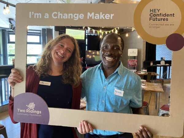 Two adults smiling and holding a cardboard frame. The frame reads "I'm a change maker"