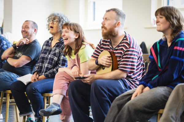 Five adults sat on chairs close together, facing towards the left. They are reacting to something off camera, with most of the laughing.