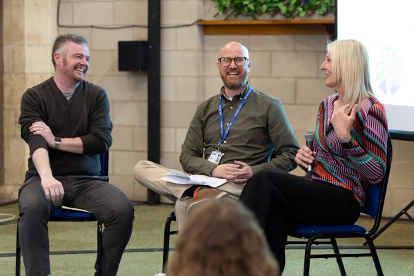 Three adults seated on chairs on a stage, deep in conversation.