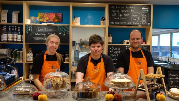 Three people standing behind a counter. They are wearing bright orange aprons and smiling at the camera.
