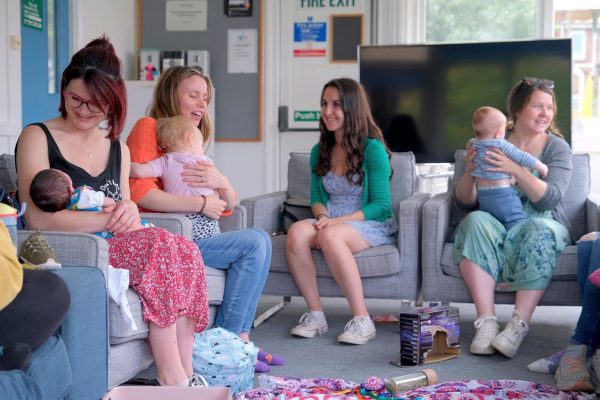 A group of four women sitting on a row of grey armchairs, each holding a baby.