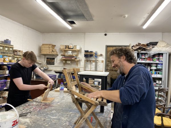 A workshop with shelves of paint and tools in the background. On the left of the photo is a boy wearing a dark t-shirt leaning over the bench working on a chair. On the right, is an older man also working on a wooden chair.