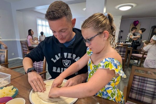 An adult and young girl working together at a table. It look like they are rolling out pizza dough.